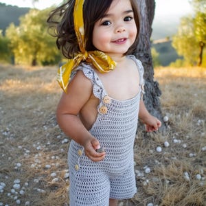 Pode incluir: Uma criança pequena vestindo um vestido de macramê cinza com uma faixa de cabeça floral amarela. O vestido tem um corpete e alças de macramê com botões de madeira. A criança está em pé em um campo de grama seca.