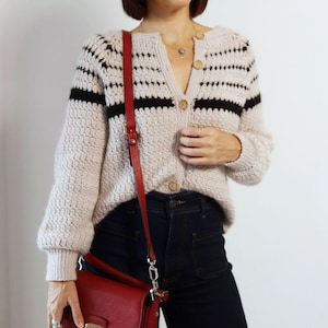May include: A beige, button-down cardigan with black stripes, paired with dark blue jeans and a red crossbody bag. The bag has a gold clasp and a red strap. The cardigan has wooden buttons.