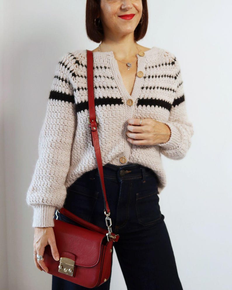May include: A beige, button-down cardigan with black stripes, paired with dark blue jeans and a red crossbody bag. The bag has a gold clasp and a red strap. The cardigan has wooden buttons.