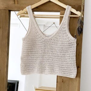 K&ouml;nnte beinhalten: Cremefarbenes, handgeh&auml;keltes Tanktop mit V-Ausschnitt, das an einem Holzb&uuml;gel h&auml;ngt. Das Oberteil hat ein strukturiertes Aussehen und wird vor einer Holzt&uuml;r und einem Spiegel pr&auml;sentiert.