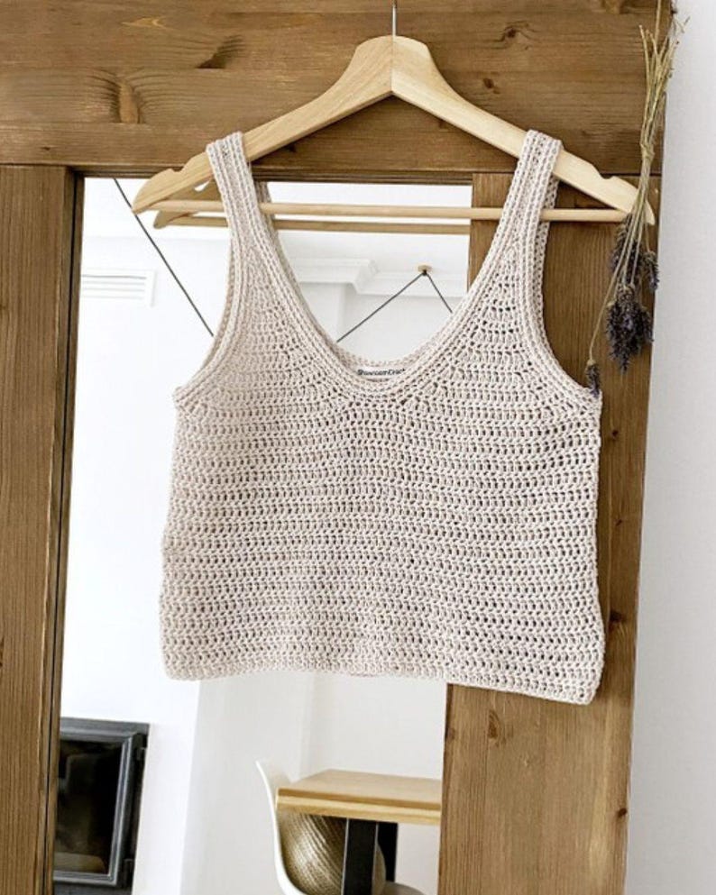K&ouml;nnte beinhalten: Cremefarbenes, handgeh&auml;keltes Tanktop mit V-Ausschnitt, das an einem Holzb&uuml;gel h&auml;ngt. Das Oberteil hat ein strukturiertes Aussehen und wird vor einer Holzt&uuml;r und einem Spiegel pr&auml;sentiert.
