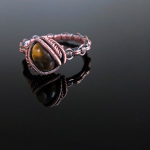 May include: A handcrafted copper wire ring with a tiger's eye stone. The ring has intricate wire wrapping around the stone and band, creating a unique design. The stone is a warm brown colour with golden streaks. The ring is displayed on a reflective black surface.