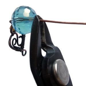 Peut inclure: Gros plan sur une pince coupante noire tenant une perle de verre facet&eacute;e bleu clair avec un fil de cuivre enroul&eacute; autour.