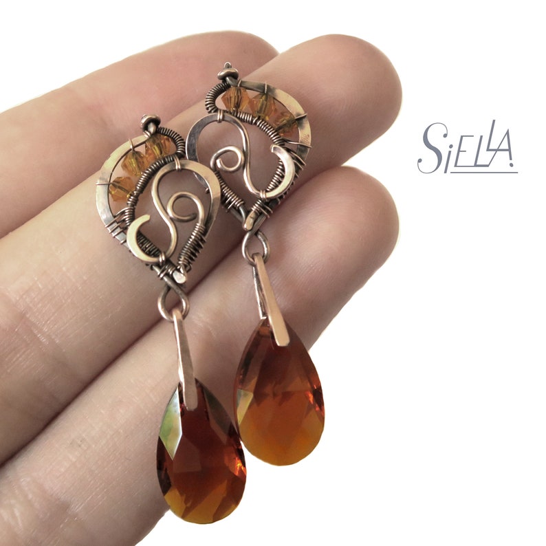 Peut inclure: Une paire de boucles d'oreilles pendantes avec un motif en forme de c&oelig;ur orn&eacute; d'un tourbillon et de perles de couleur ambre. Les boucles d'oreilles sont en fil de cuivre et ont un cristal ambre en forme de larme suspendu &agrave; chaque c&oelig;ur.