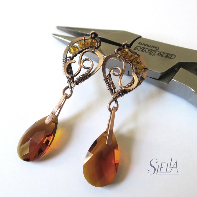 Peut inclure: Une paire de boucles d'oreilles en fil de cuivre avec des cristaux ambr&eacute;s en forme de goutte. Les boucles d'oreilles pr&eacute;sentent des motifs de filature complexes avec de petites perles dor&eacute;es.