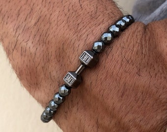 Pode incluir: Um bracelete de contas pretas com um fecho de halteres prateado. As contas são facetadas e têm um acabamento fosco.
