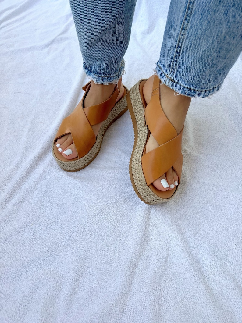 May include: Tan leather sandals with a crisscross strap design. The sandals have a woven jute platform sole. The image shows the sandals being worn with blue jeans.
