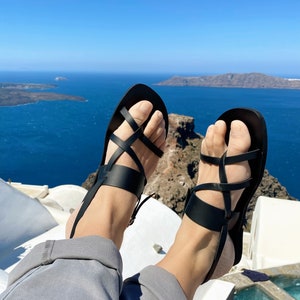 May include: A pair of black leather sandals with criss-cross straps. The sandals are worn by a person sitting on a white surface with a view of the ocean in the background.