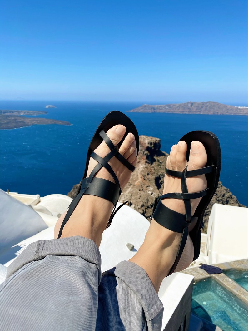 May include: A pair of black leather sandals with criss-cross straps. The sandals are worn by a person sitting on a white surface with a view of the ocean in the background.