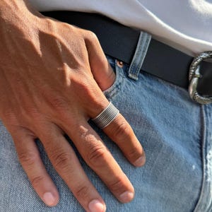 May include: A silver ring with a stacked, ridged design. The ring is worn on a finger, set against a backdrop of blue denim jeans, a black belt, and a white t-shirt.