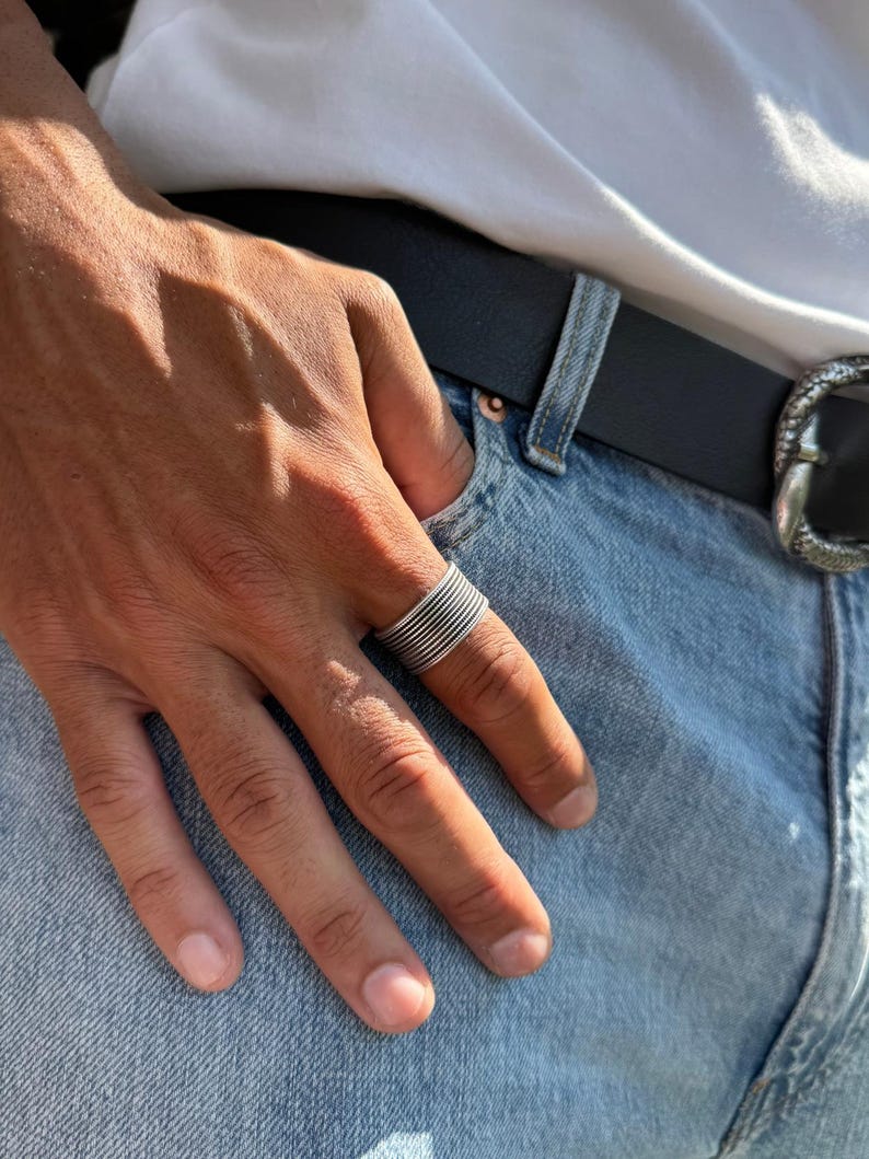 May include: A silver ring with a stacked, ridged design. The ring is worn on a finger, set against a backdrop of blue denim jeans, a black belt, and a white t-shirt.