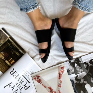 May include: A pair of black leather double strap sandals with a simple design. The sandals are worn by a person with bare feet and jeans.