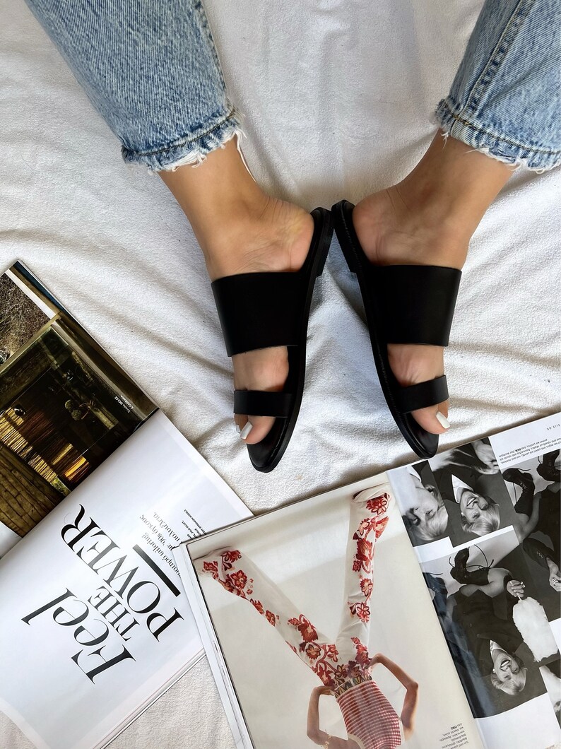 May include: A pair of black leather double strap sandals with a simple design. The sandals are worn by a person with bare feet and jeans.