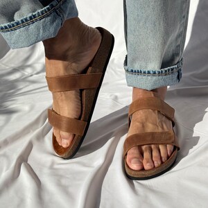 May include: Brown leather sandals with two adjustable straps and a cork footbed. The sandals are shown with light blue denim jeans. The sandals are designed for comfort and casual wear.
