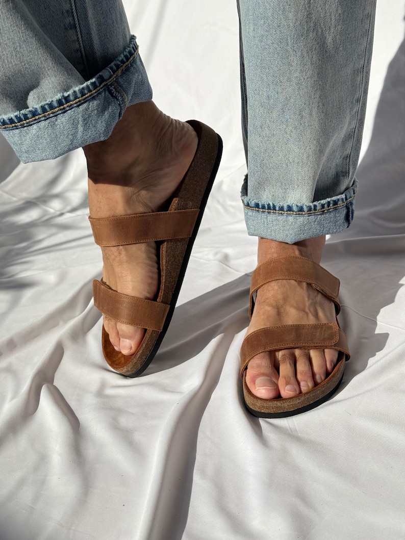 May include: Brown leather sandals with two adjustable straps and a cork footbed. The sandals are shown with light blue denim jeans. The sandals are designed for comfort and casual wear.