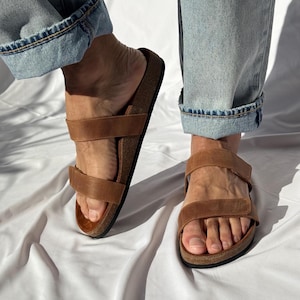 May include: Brown leather sandals with two adjustable straps. The sandals have a cork footbed and are worn with blue jeans. The sandals are a neutral color and can be worn with many outfits.