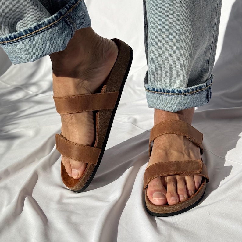May include: Brown leather sandals with two adjustable straps. The sandals have a cork footbed and are worn with blue jeans. The sandals are a neutral color and can be worn with many outfits.