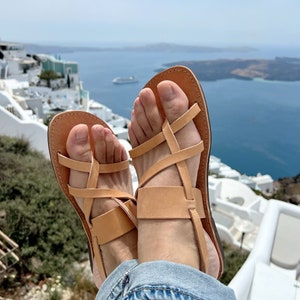 May include: A pair of tan leather sandals with criss-cross straps. The sandals are worn by a person sitting on a white surface with a view of a blue ocean and white buildings in the background.