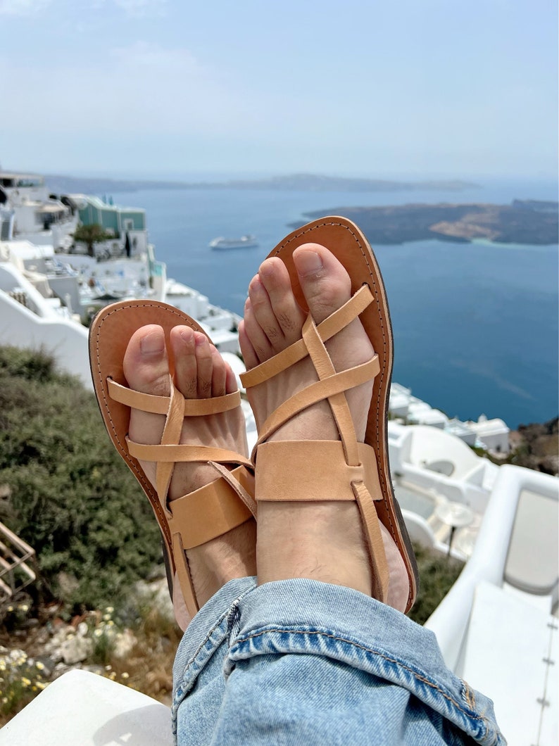 May include: A pair of tan leather sandals with criss-cross straps. The sandals are worn by a person sitting on a white surface with a view of a blue ocean and white buildings in the background.