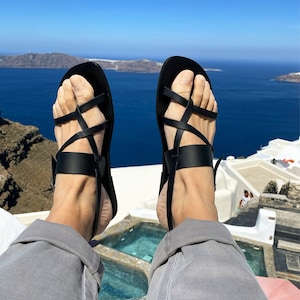 May include: A pair of black leather sandals with criss-cross straps. The sandals are worn by a person sitting on a white ledge overlooking a body of water.