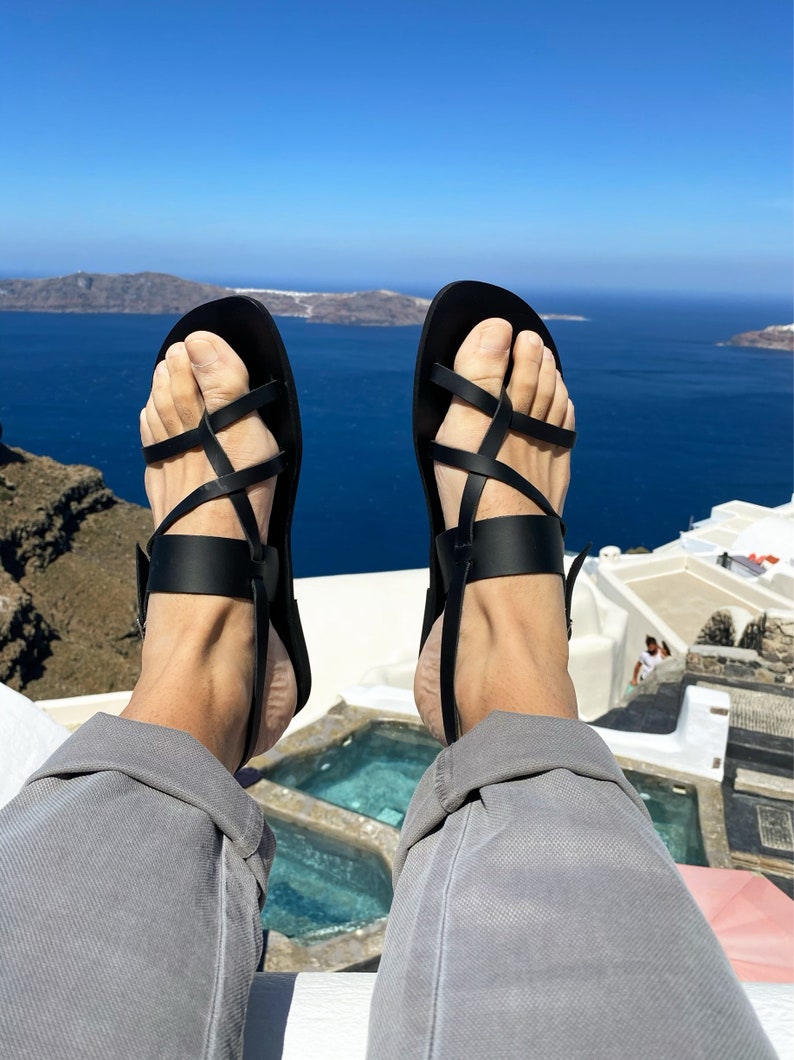 May include: A pair of black leather sandals with criss-cross straps. The sandals are worn by a person sitting on a white ledge overlooking a body of water.