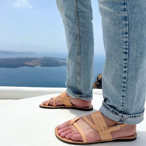 May include: A pair of tan leather sandals with a criss-cross strap design. The sandals are worn by a person standing on a white surface with a view of a blue body of water in the background.