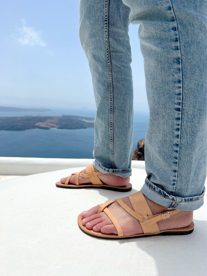 May include: A pair of tan leather sandals with a criss-cross strap design. The sandals are worn by a person standing on a white surface with a view of a blue body of water in the background.