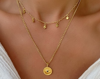 Könnte beinhalten: Eine goldene Kettenkette mit einem kleinen Augenanhänger und einer zarten Kette mit kleinen quadratischen Anhängern. Die Kette ist geschichtet und hat ein schlichtes, elegantes Design.