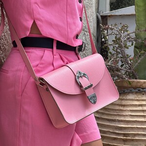 Op de afbeelding: Een roze leren schoudertas met een zilveren gespsluiting. De tas wordt gedragen over een roze outfit.