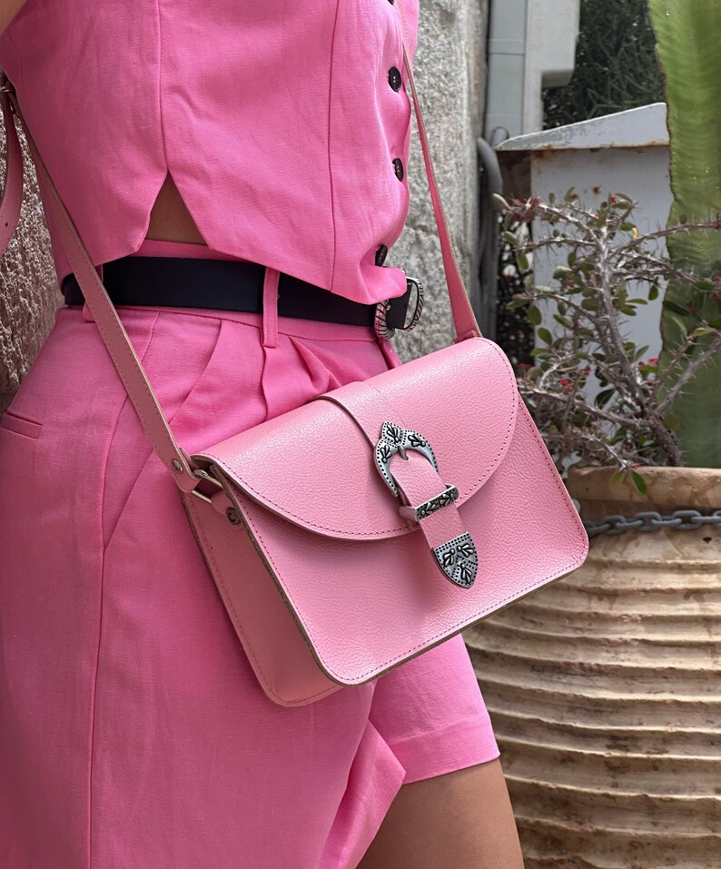 Op de afbeelding: Een roze leren schoudertas met een zilveren gespsluiting. De tas wordt gedragen over een roze outfit.