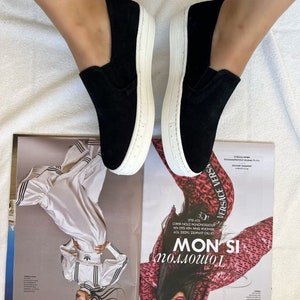 May include: A pair of black slip-on canvas shoes with white soles. The shoes are worn by a person with bare legs and blue jeans.