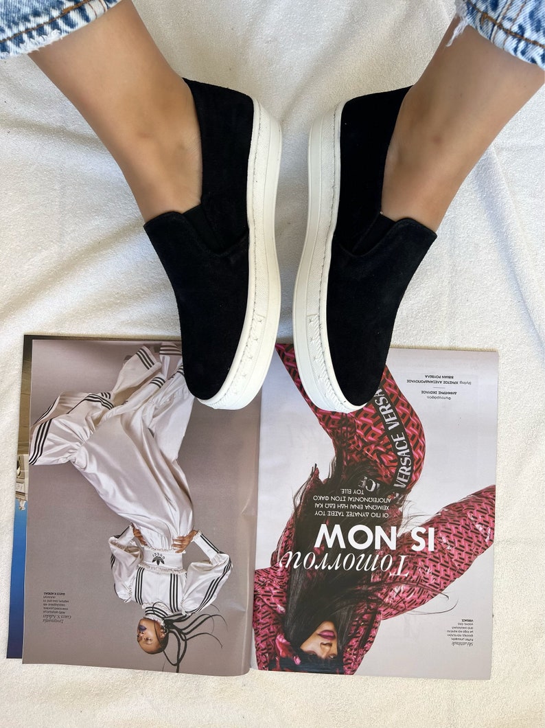 May include: A pair of black slip-on canvas shoes with white soles. The shoes are worn by a person with bare legs and blue jeans.