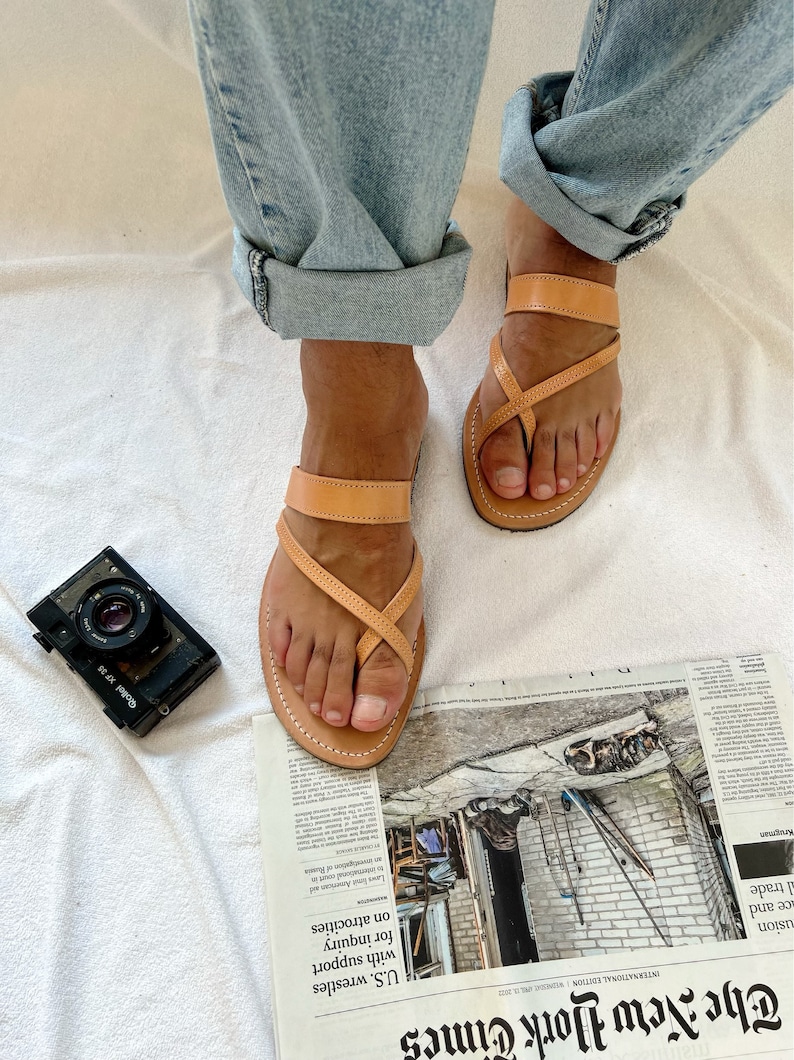 May include: A pair of tan leather sandals with a criss-cross strap design. The sandals are worn on a person's feet, with the toes visible. The sandals are on top of a newspaper with the text "The New York Times" visible.