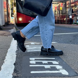 May include: A person wearing blue jeans and black lace-up shoes walks across a street with a black leather bag. The street has a white painted line and the word "LEFT" painted on the asphalt.