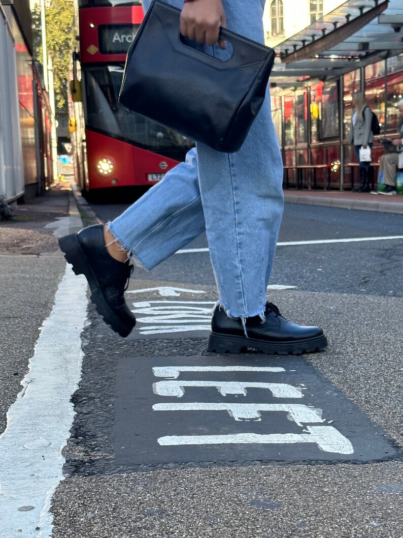 May include: A person wearing blue jeans and black lace-up shoes walks across a street with a black leather bag. The street has a white painted line and the word "LEFT" painted on the asphalt.
