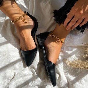 May include: Black pointed-toe flats with a gold chain ankle bracelet. The shoes have a low heel and a sleek, minimalist design. The gold chain adds a touch of elegance to the simple, classic style. The flats are shown on a white surface.