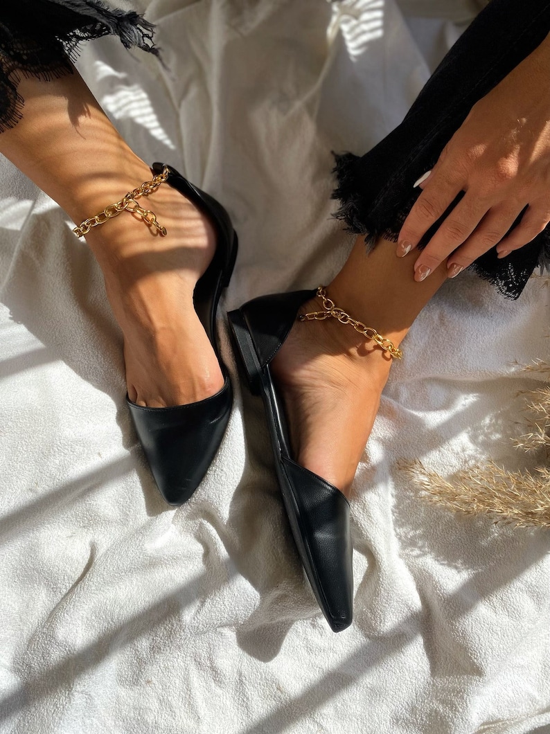 May include: Black pointed-toe flats with a gold chain ankle bracelet. The shoes have a low heel and a sleek, minimalist design. The gold chain adds a touch of elegance to the simple, classic style. The flats are shown on a white surface.