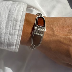 Può includere: Bracciale in argento con due intarsi rettangolari in pietra marrone e beige. Il bracciale ha un design a catena e viene indossato su un polso. Lo sfondo è in tessuto bianco.
