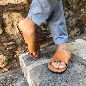 May include: A pair of brown leather sandals with a criss-cross strap design. The sandals are worn by a person wearing light blue pants.