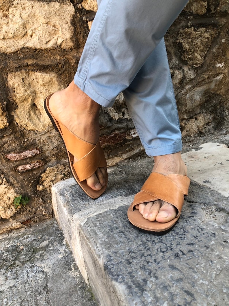 May include: A pair of brown leather sandals with a criss-cross strap design. The sandals are worn by a person wearing light blue pants.