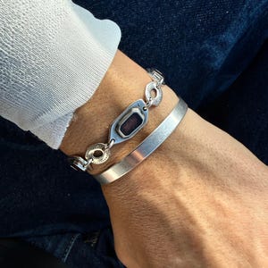 May include: Two silver bracelets are shown, one with an oblong, dark-colored stone and the other a simple band. The bracelets are worn on a wrist, with a white long-sleeved shirt and blue jeans in the background.
