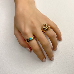 May include: Two gold rings on a hand. One ring has a turquoise stone and the other ring has an eye design.