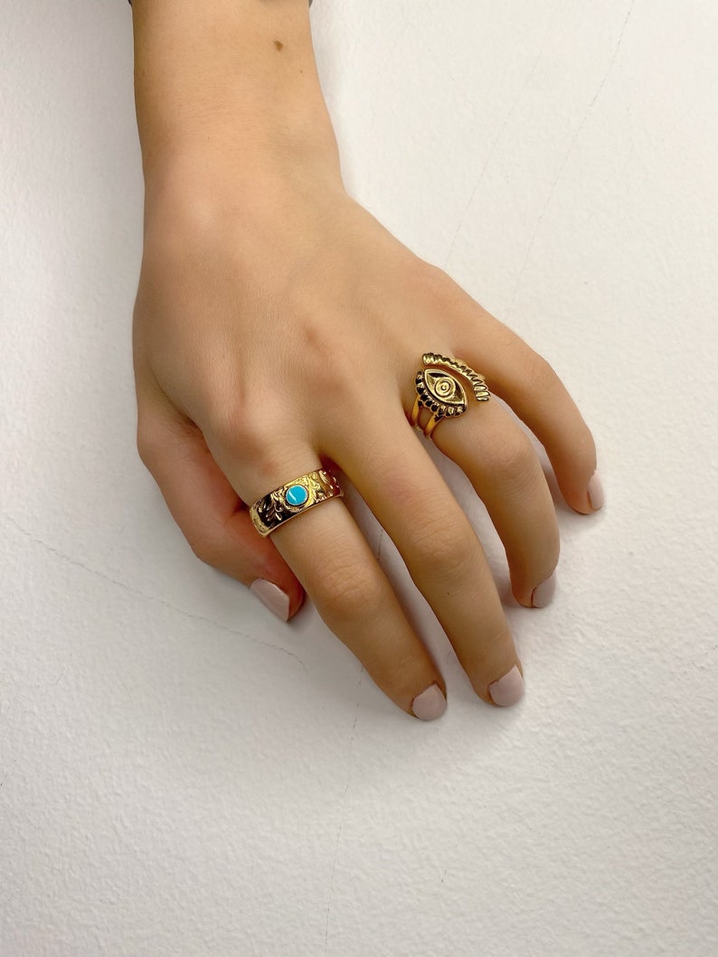 May include: Two gold rings on a hand. One ring has a turquoise stone and the other ring has an eye design.