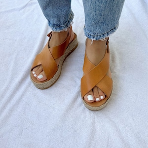 May include: Tan leather sandals with a crisscross design and a woven jute platform sole. The sandals have a buckle closure and are worn with blue jeans. The sandals are a neutral color and are suitable for casual wear.