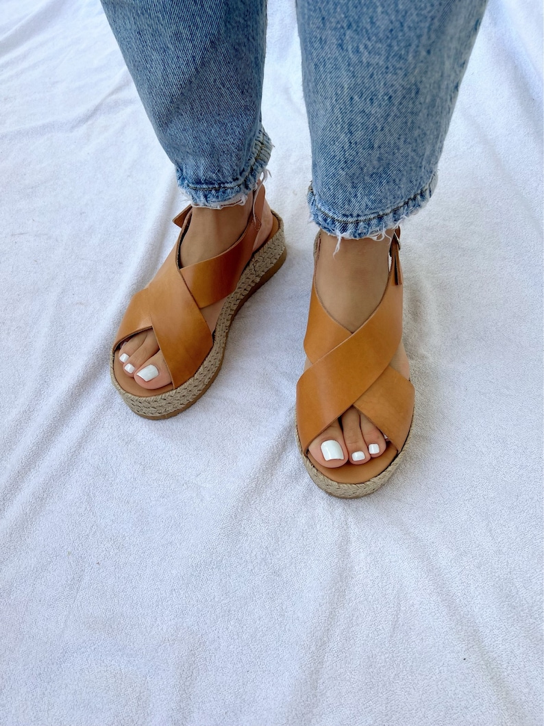 May include: Tan leather sandals with a crisscross design and a woven jute platform sole. The sandals have a buckle closure and are worn with blue jeans. The sandals are a neutral color and are suitable for casual wear.