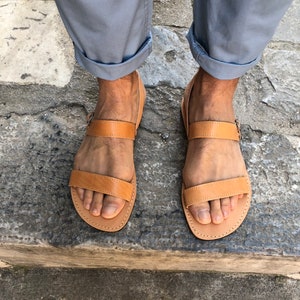 May include: A pair of brown leather sandals with two straps and a buckle. The sandals are worn by a person with rolled up pants.