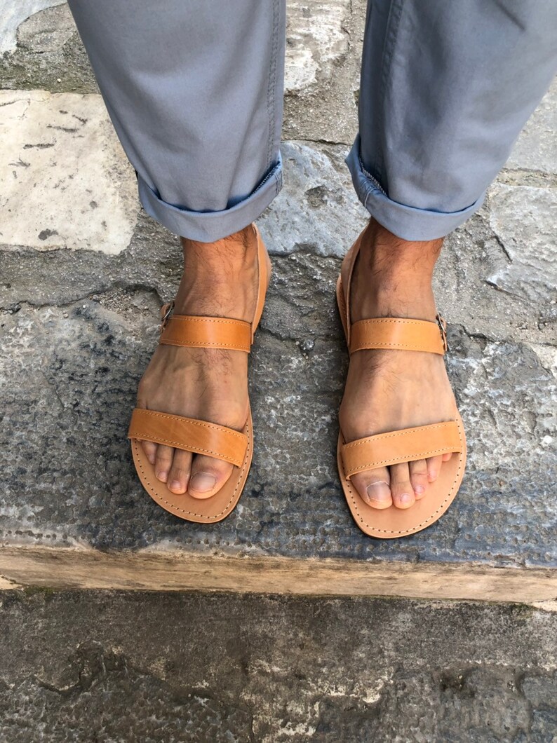 May include: A pair of brown leather sandals with two straps and a buckle. The sandals are worn by a person with rolled up pants.