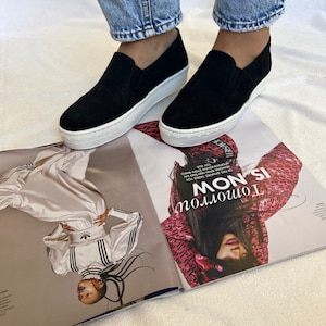 May include: A pair of black suede platform slip-on shoes with white soles. The shoes are worn by a person wearing blue jeans.