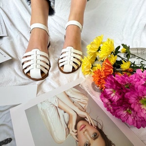 Puede incluir: Sandalias blancas de cuero con un dise&ntilde;o enjaulado y cierre de hebilla. Las sandalias est&aacute;n sobre una superficie blanca con un ramo de flores.