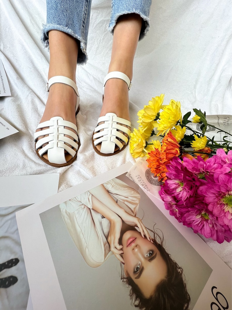 Puede incluir: Sandalias blancas de cuero con un dise&ntilde;o enjaulado y cierre de hebilla. Las sandalias est&aacute;n sobre una superficie blanca con un ramo de flores.